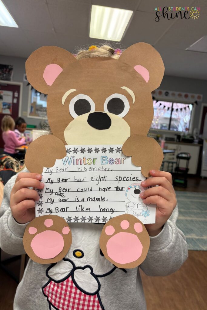 A young student holding a Winter Bear craftivity, featuring a large paper bear with pink paws attached to a writing prompt about bears. The writing includes fun facts about bear species, hibernation, and diet. This engaging winter craftivity combines creativity and learning, making it a great seasonal classroom activity.