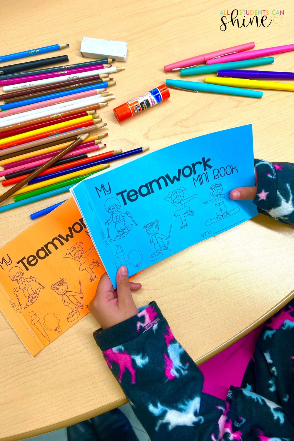 Winter sports-themed teamwork mini-book activity for elementary students. A child holds a blue 'My Teamwork Mini Book' while an orange version rests on the table. The workspace is filled with colorful pencils, markers, an eraser, and a glue stick. A fun social-emotional learning activity promoting collaboration in the classroom.