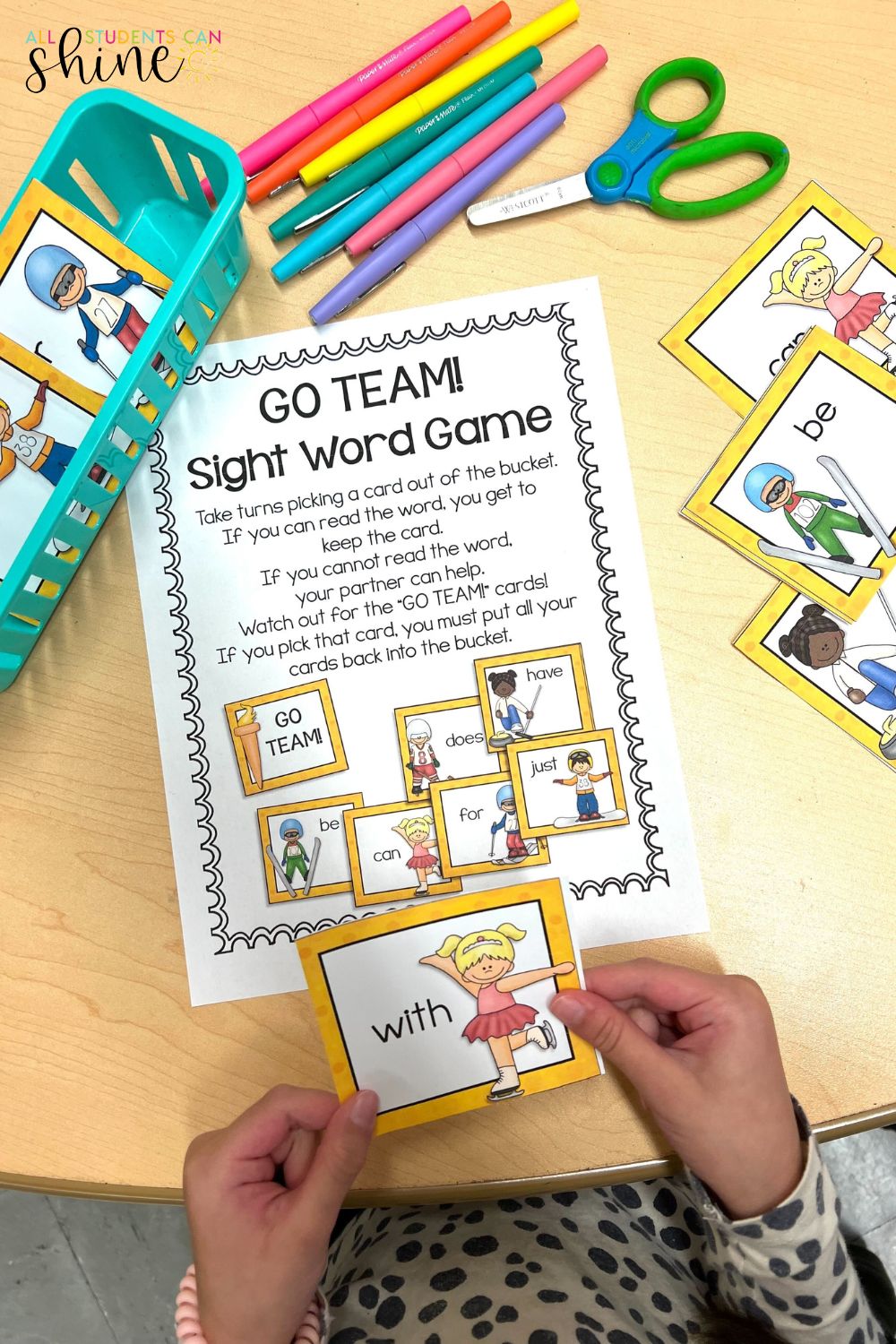 Winter sports-themed sight word game for early learners. A child holds a sight word card featuring an ice skater while playing the 'GO TEAM!' Sight Word Game. The table includes a worksheet with game instructions, a blue basket filled with word cards, colorful markers, and scissors. A fun literacy center activity for kindergarten and first grade.