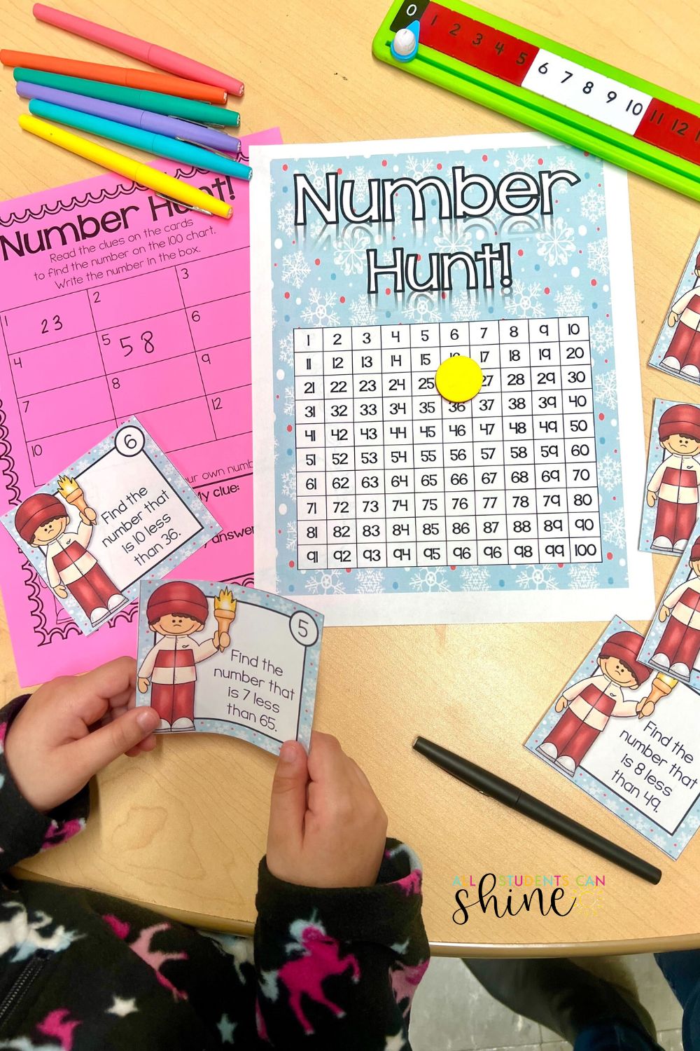 Winter sports-themed number hunt activity for K-1 students. A child wearing a cozy, patterned fleece holds a math clue card while working on a 100-chart number search. The table includes a pink worksheet, colorful markers, a number line, and additional math clue cards featuring a winter athlete. Engaging math center for early learners.