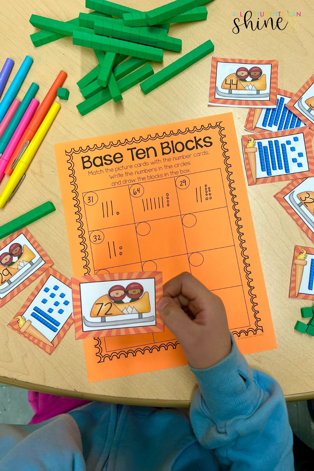 Winter sports-themed base ten blocks math activity for elementary students. A child holds a number card featuring a bobsled, matching it to numbers on an orange worksheet. Green base ten blocks, colorful markers, and additional number cards with winter sports illustrations are on the table. A hands-on learning activity for place value practice.